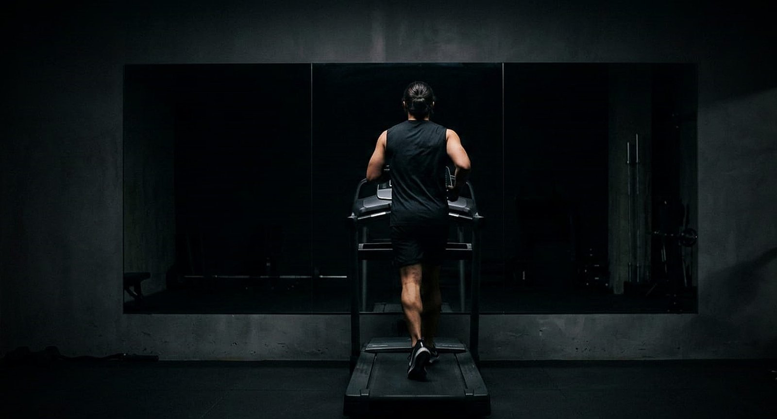 Pessoa vista de costas correndo em esteira diante de espelho que não reflete sua imagem.